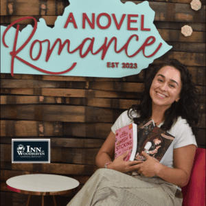 Woman with long black hair, a white t-shirt, and khaki pants sits in a chair holding two of her books. In the background is a rustic wood wall and a light blue cut-out of the state of Kentucky that says "A Novel Romance"