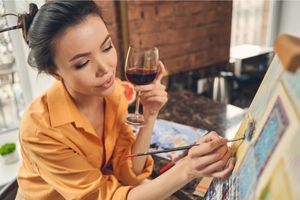 Woman in a peach blouse is holding a glass of red wine and painting on a canvas.