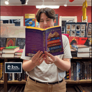 Person with short brown hair, a white polo, and khaki pants reads a purple-covered book while standing in front of bookshelves.