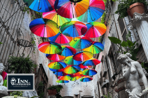 A narrow alleyway adorned with dozens of hanging rainbow-colored umbrellas, surrounded by greenery and classical statues. The whimsical overhead display creates a magical, art-filled urban escape.