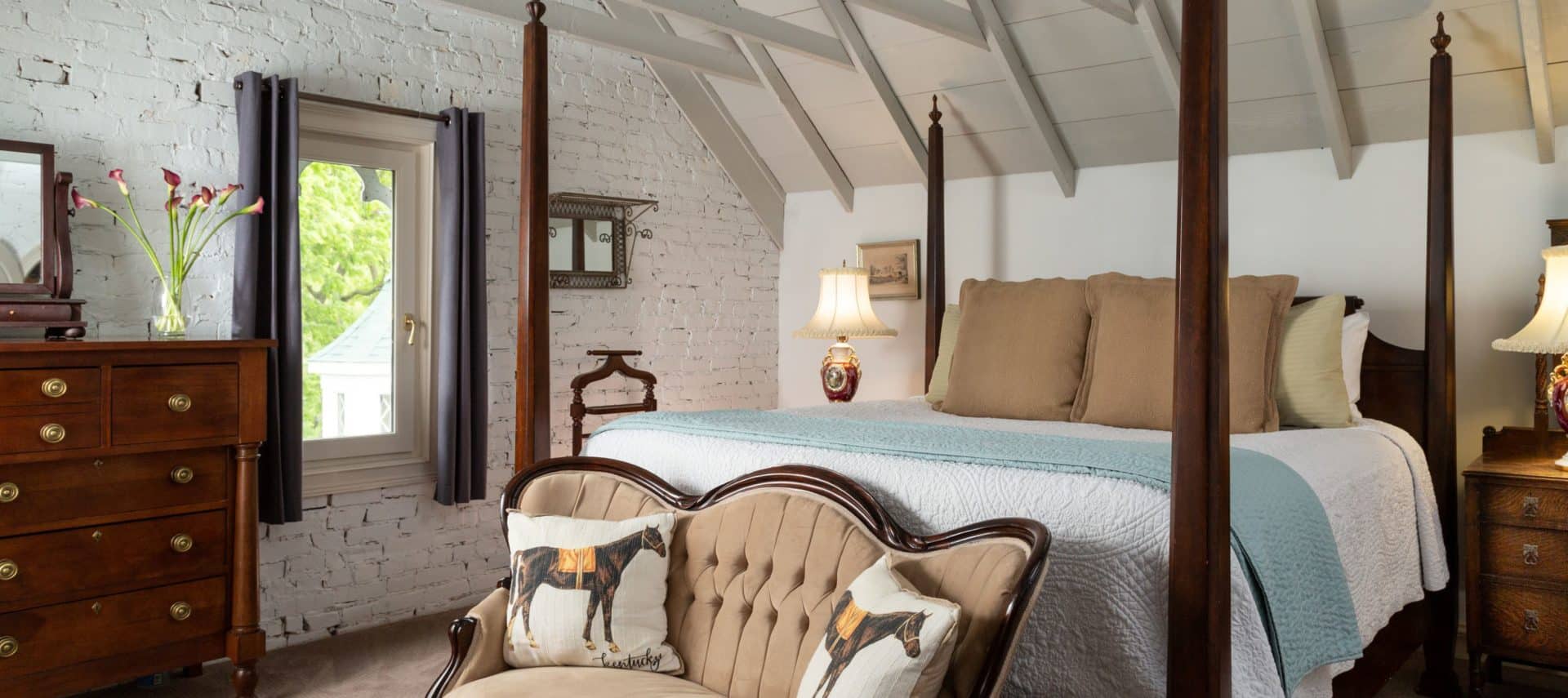 Very large room with vaulted ceilings. A king four-post bed with white bedspread and decorative pillows is on the right of the image with a beige loveseat at the foot of the bed also with decorative pillows. A window in the middle of the picture and a dresser on the left side.