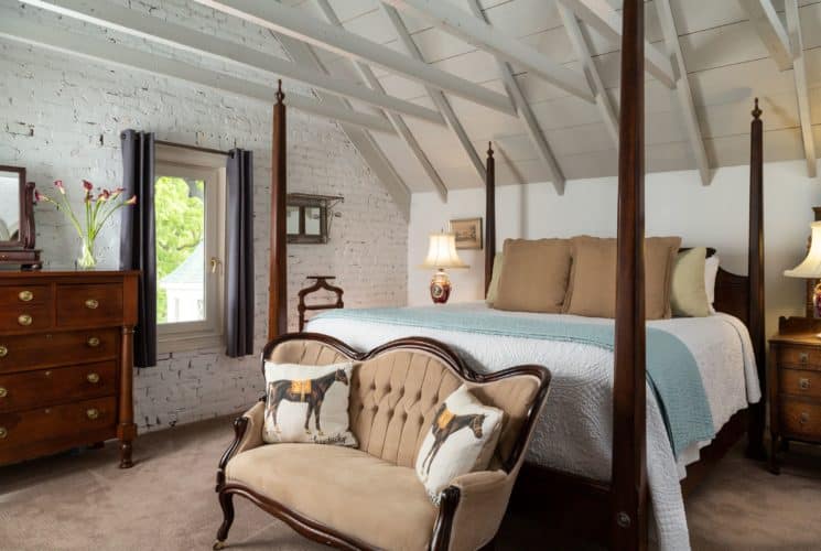 Very large room with vaulted ceilings. A king four-post bed with white bedspread and decorative pillows is on the right of the image with a beige loveseat at the foot of the bed also with decorative pillows. A window in the middle of the picture and a dresser on the left side.