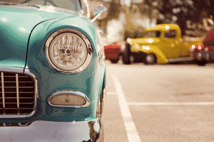 The front left headlight of a light blue vintage car with other vintages cars parked in the background.