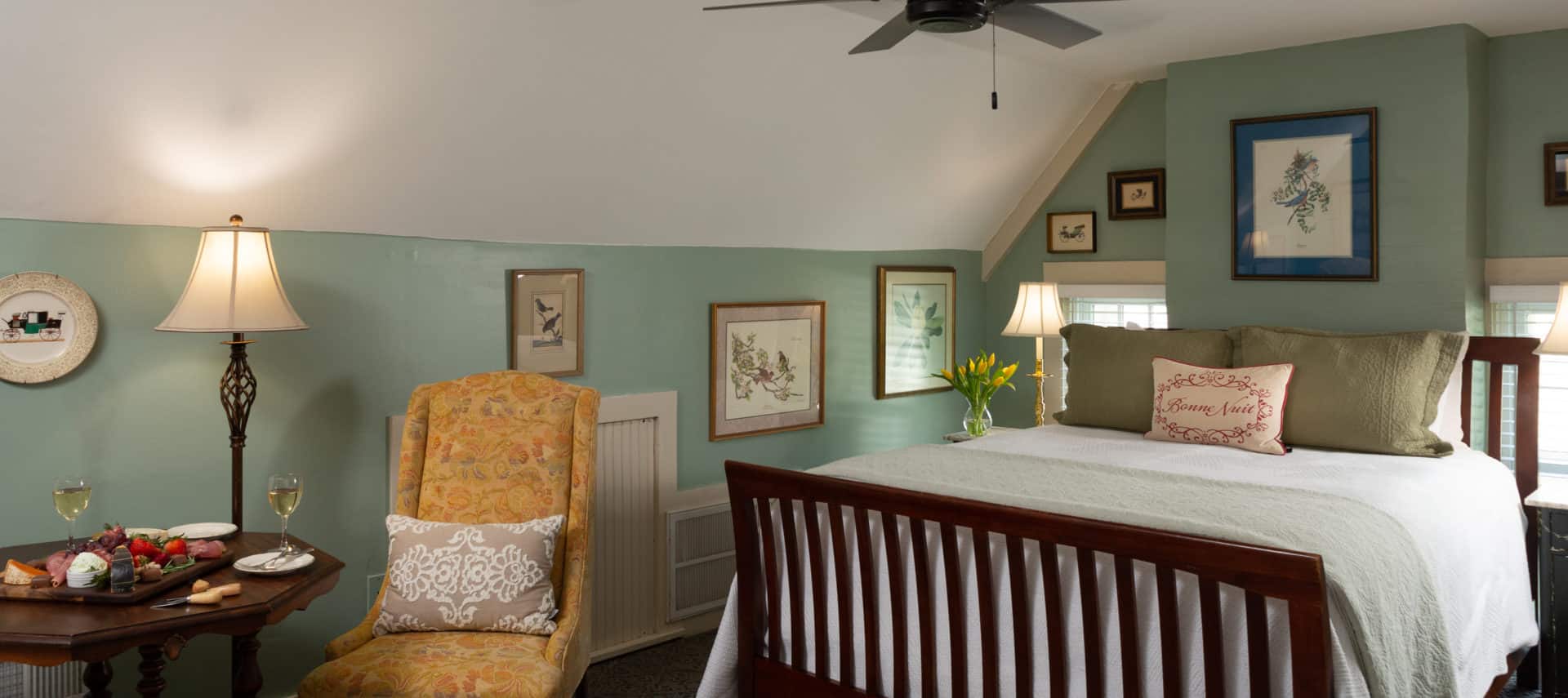 queen sized sleigh bed with white and green linins and sitting area with two chairs and a lamp and a snack board setting with two glasses of whine. Pale green walls and an arrangement of yellow tulips next to the bed