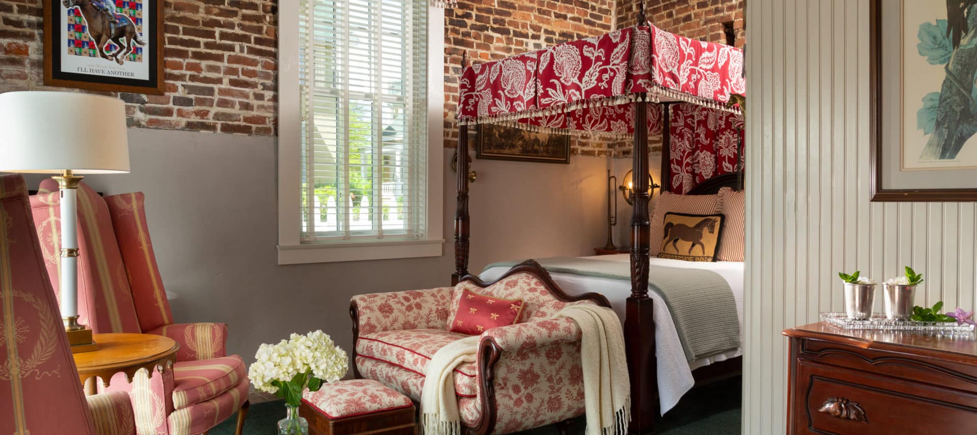 queen sized four-post bed with red and white canopy and matching valance on a large window. Brick walls and a love seat and foot stool. Side table with books and an arrangement of white flowers