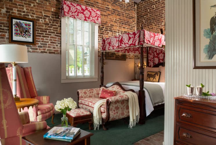 queen sized four-post bed with red and white canopy and matching valance on a large window. Brick walls and a love seat and foot stool. Side table with books and an arrangement of white flowers