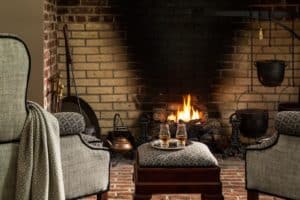 A gray chair in front of a fireplace with an ottoman and two glasses of bourbon on a tray in front of the fire.