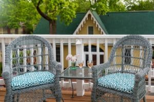 A patio area with two wicker chairs in front of a white rail. Image of a yellow carriage house seen through the railing between the chairs.