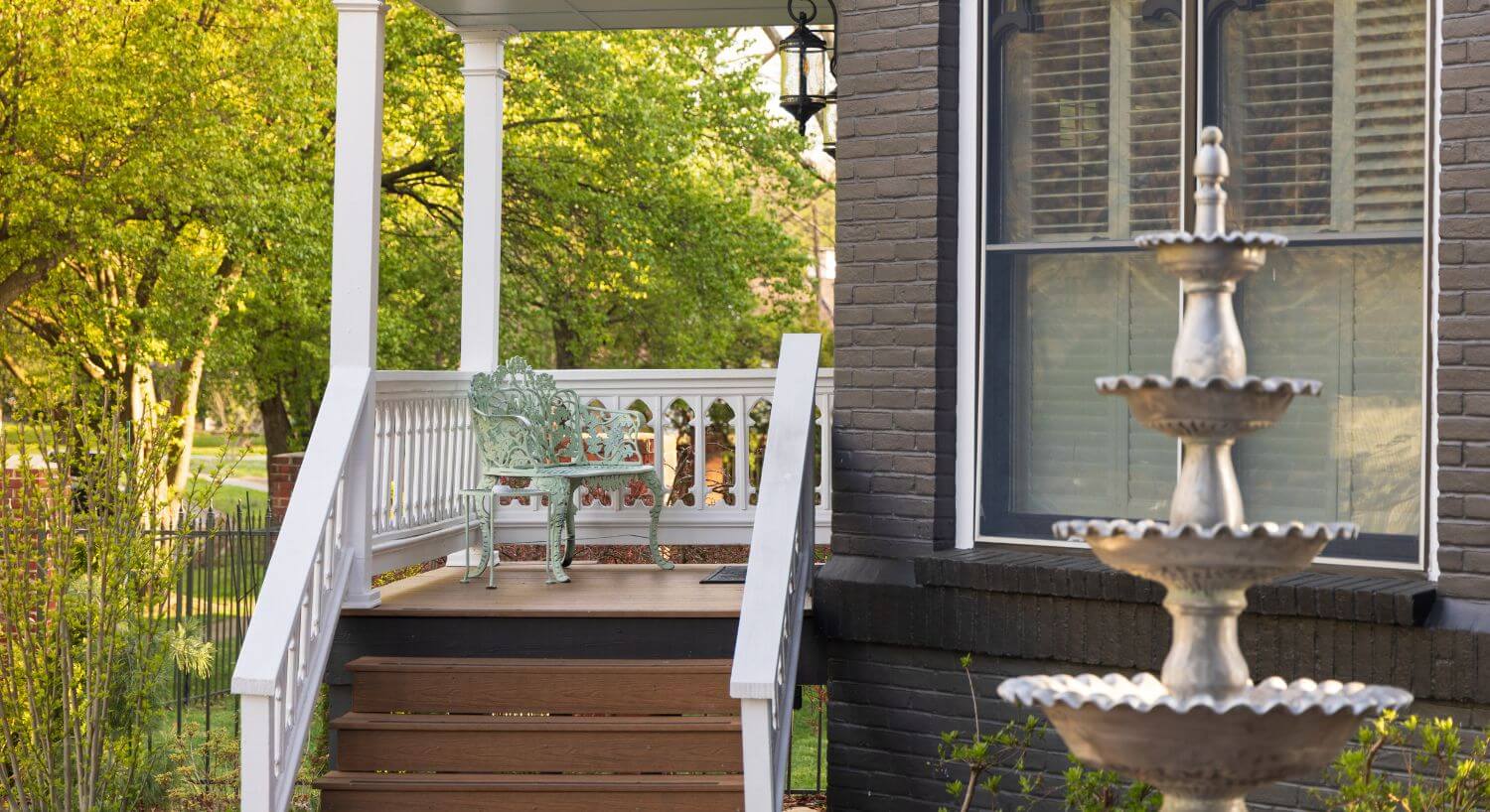 stairs up to small porch with mint green metal seat, four-tier fountain in front of window on gray house to right of porch