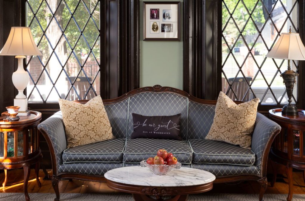 A blue sofa with decorative pillows sits in front of two very large gothic style windows and a marble coffee table sits in front of the sofa with a bowl of red apples.