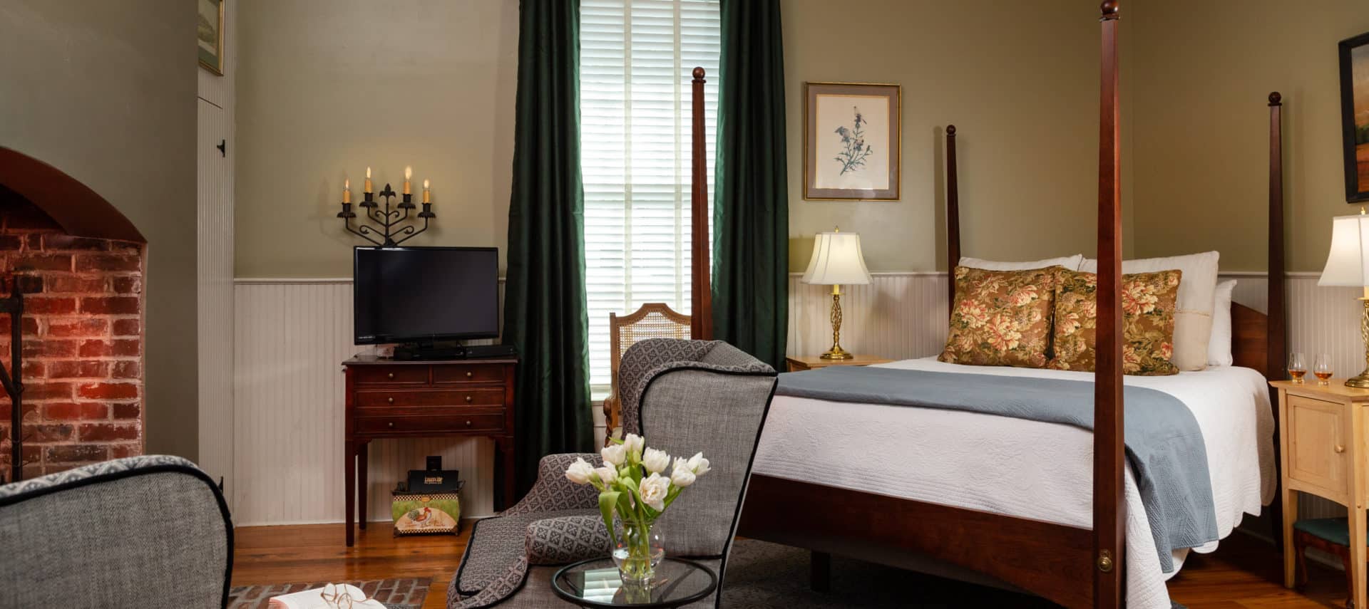 Earth-toned walls with a large fireplace to the left of the image. Two large grey chairs with an arrangement of white flowers in front of the fireplace and a queen sized bed with white bedspread and decorative pillows. A large window in the background with dark evergreen drapes