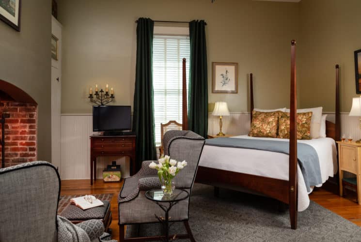 Earth-toned walls with a large fireplace to the left of the image. Two large grey chairs with an arrangement of white flowers in front of the fireplace and a queen sized bed with white bedspread and decorative pillows. A large window in the background with dark evergreen drapes