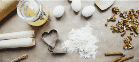 Eggs, flour, two silver heart cookie cutters, nuts, cinnamon, and a jar are scattered across a baking table.