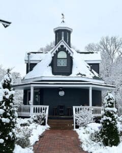 A dark gray Victorian-style house with white trim sits under fresh snowfall, featuring a wraparound porch, brick walkway, and a small cupola on the roof, surrounded by snow-covered trees and shrubs.