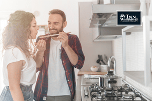 Man and woman standing in front of a stove, with him serving her a wooden spoonful of food.