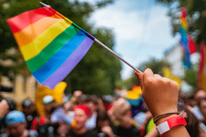 Hand holding the traditional rainbow Pride flag in the foreground with people celebrating in the background.