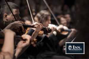 One male violinist with shoulder length black hair plays his instruments surrounded by other violinists in an orchestra.