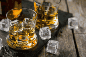 Two glasses of bourbon with cubed ice on a wooden table.