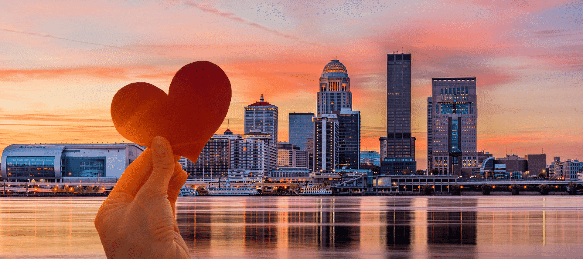 Louisville Kentucky city skyline with a pink and orange sunset and a hand holding a cutout paper heart in the foreground.