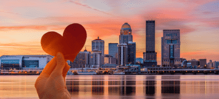 Louisville Kentucky city skyline with a pink and orange sunset and a hand holding a cutout paper heart in the foreground.