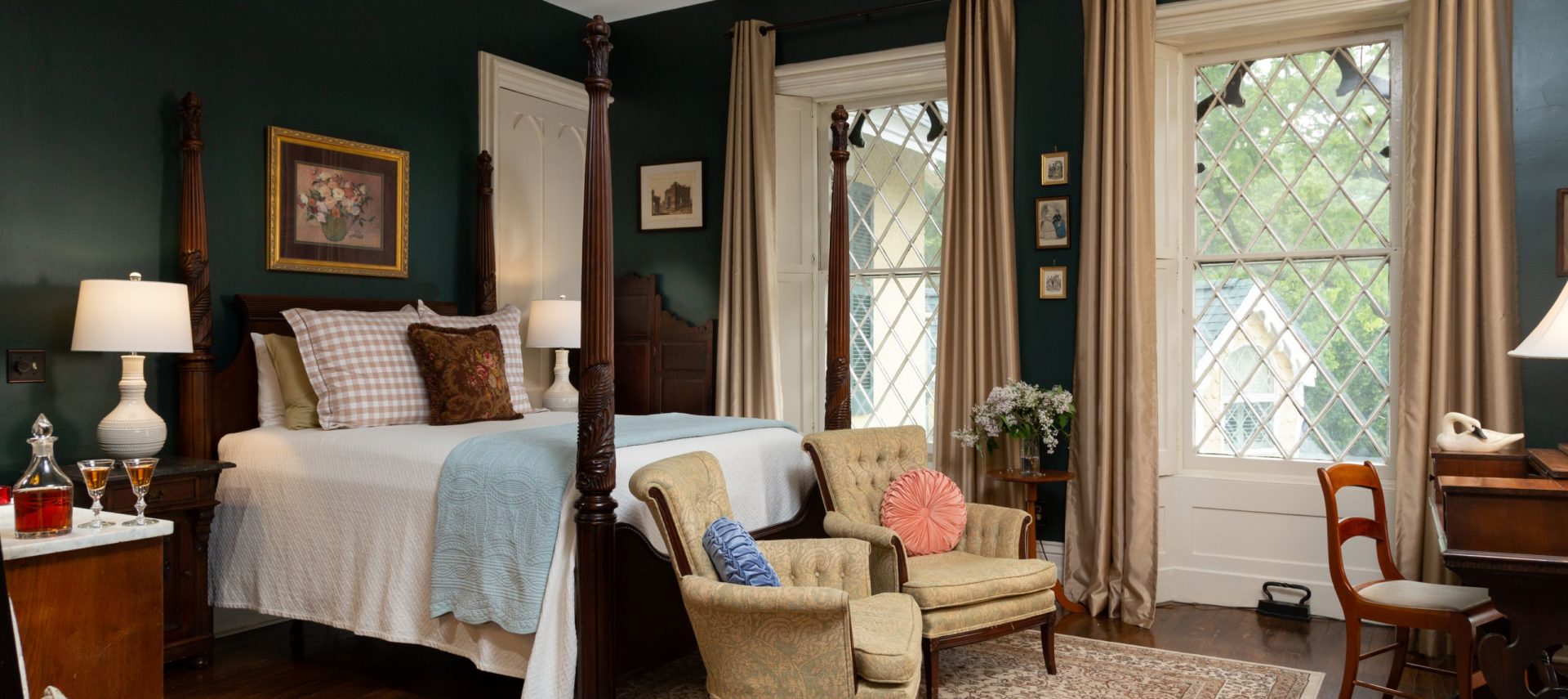 Spacious room with dark evergreen painted walls and two gothic style windows. A queen four-post bed in the center of the room with a white bedspread and decorative pillows. Two chairs sit at the foot of the bed on a large decorative area rug.