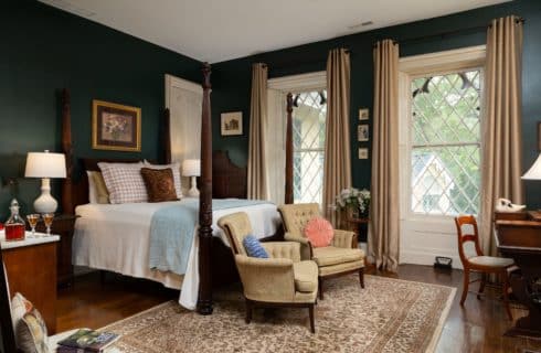 Spacious room with dark evergreen painted walls and two gothic style windows. A queen four-post bed in the center of the room with a white bedspread and decorative pillows. Two chairs sit at the foot of the bed on a large decorative area rug.