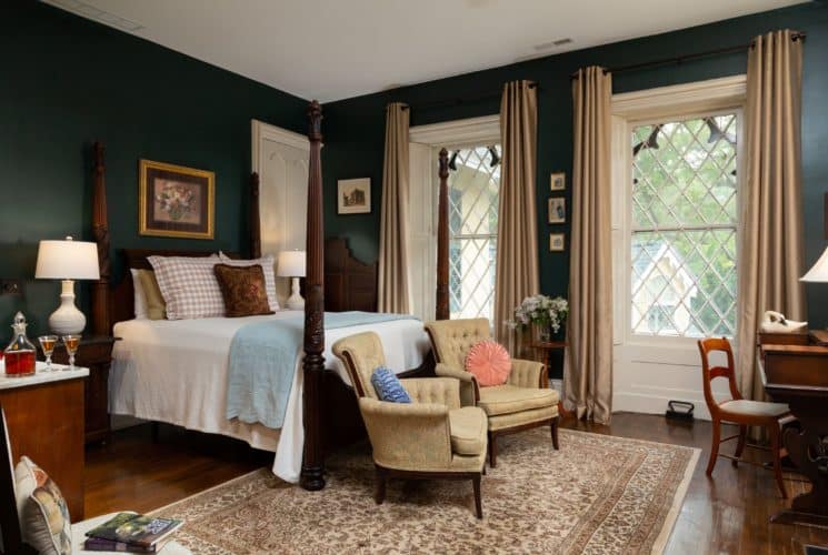 Spacious room with dark evergreen painted walls and two gothic style windows. A queen four-post bed in the center of the room with a white bedspread and decorative pillows. Two chairs sit at the foot of the bed on a large decorative area rug.