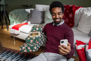 Black man in a maroon sweater holds a present and takes a selfie with his phone.
