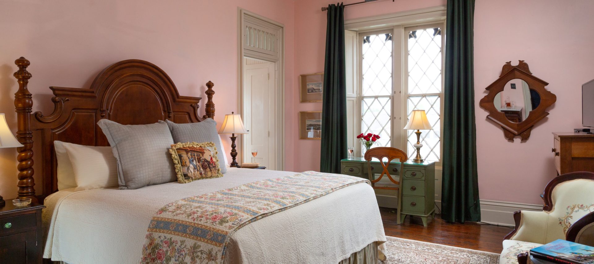 Large room with light pink walls and a large gothic style window with dark drapes. A king bed with white bedspread and decorative pillows with a large wood headboard is on the left side of the image and a writing desk is under the windows. Two chairs are on the right side of the picture