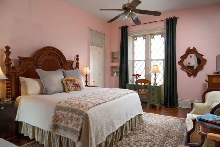 Large room with light pink walls and a large gothic style window with dark drapes. A king bed with white bedspread and decorative pillows with a large wood headboard is on the left side of the image and a writing desk is under the windows. Two chairs are on the right side of the picture