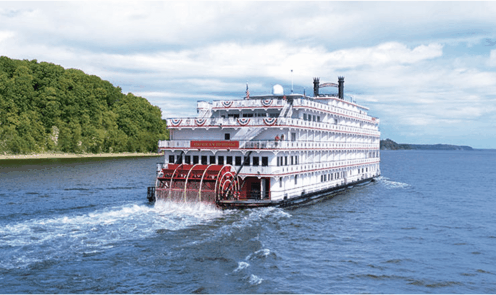 White, multi-deck riverboat with a red paddle wheel, cruising on a blue river beside a tree-lined shore.