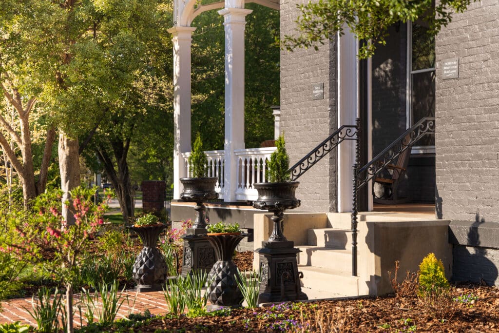 Brick house porch with black wrought-iron railing, steps, and decorative planters in a blooming garden setting.