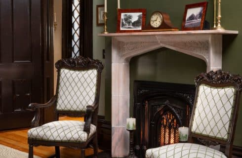 Antique chairs in front of a lit fireplace and old pictures on the mantle.