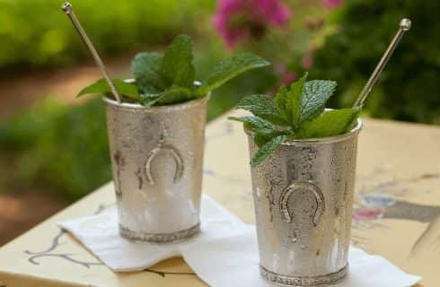 Two silver metal cups with metal stir sticks and mint sprigs sitting on white napkins on a yellow table