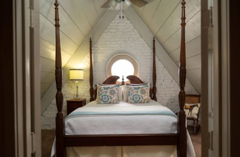 Image is of the second bedroom in the suite. A queen sized four-post bed is the center with a round window above the headboard. Vaulted ceilings