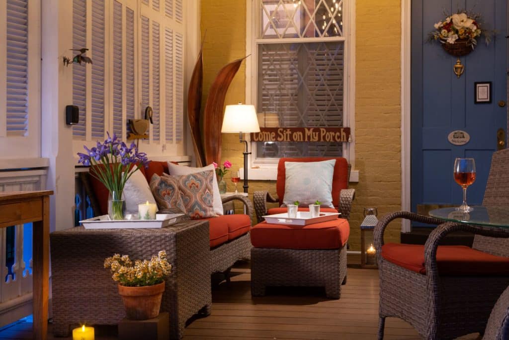 Back patio lit with Edison lights. Hellow walls with white shutters and a blue door with floral arrangement hanging on it. Red upholstered patio furniture and assorted drinks implying friends hanging out.