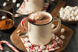 White ceramic mug of hot chocolate with a candy cane and whipped cream in it, on top of a napkin and wooden table.