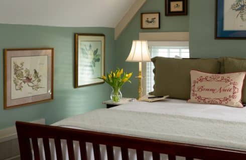 sleigh bed in the foreground with a white bedspread and decorative pillows and a journal on the bed. Background of pale green walls, framed pictures of birds and an arrangement of yellow tulips