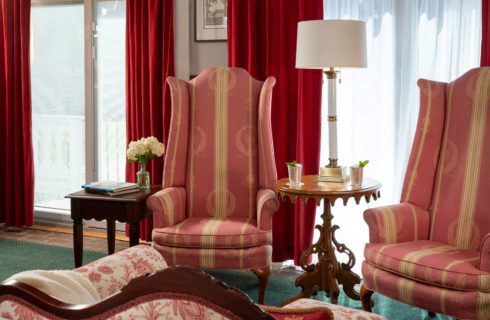 two victorian high-back pink chairs with a coffee table between with two cocktails. Large windows behind the chairs with red drapes