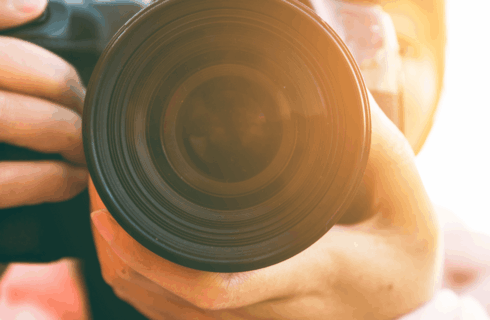 Close-up of a photographer holding a camera lens toward the viewer, with warm lighting