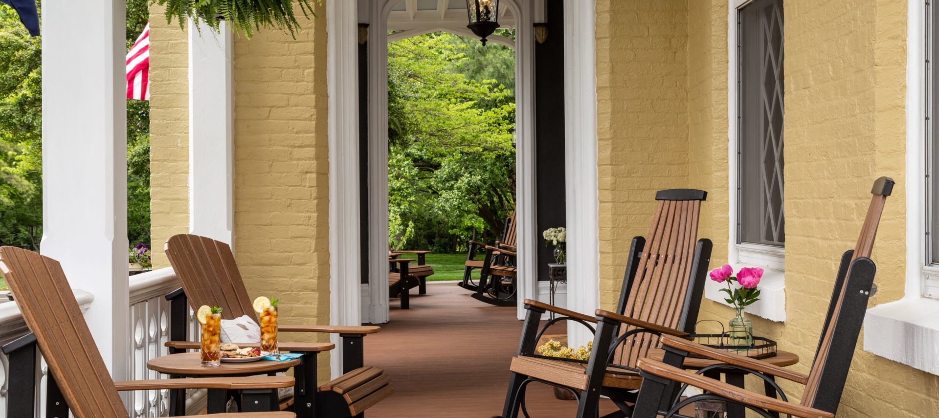 Sunny front porch with rocking and adirondack chairs. A hanging plant and a vase of pink flowers and two glasses of iced tea with lemon complete this summer themed picture.