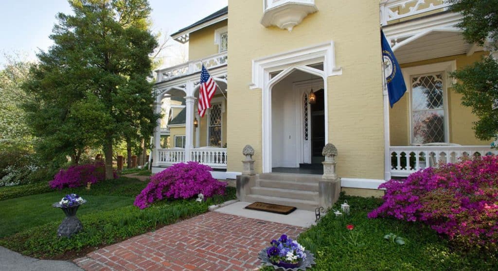 Exterior view of the property painted cream with white trim surrounded by green trees, green grass, and purple bushes