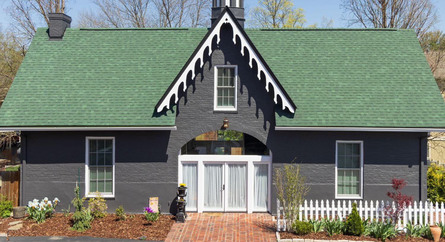 back view of gray house with green roof, garden with flowers & plants surrounded by mulch, glass double doors with white curtains
