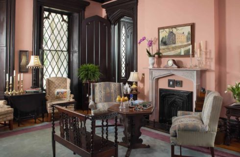Large gathering room with hardwood floors, light pink walls, large windows, upholstered chairs, dark wooden bench and table, fireplace, and large gray area rug