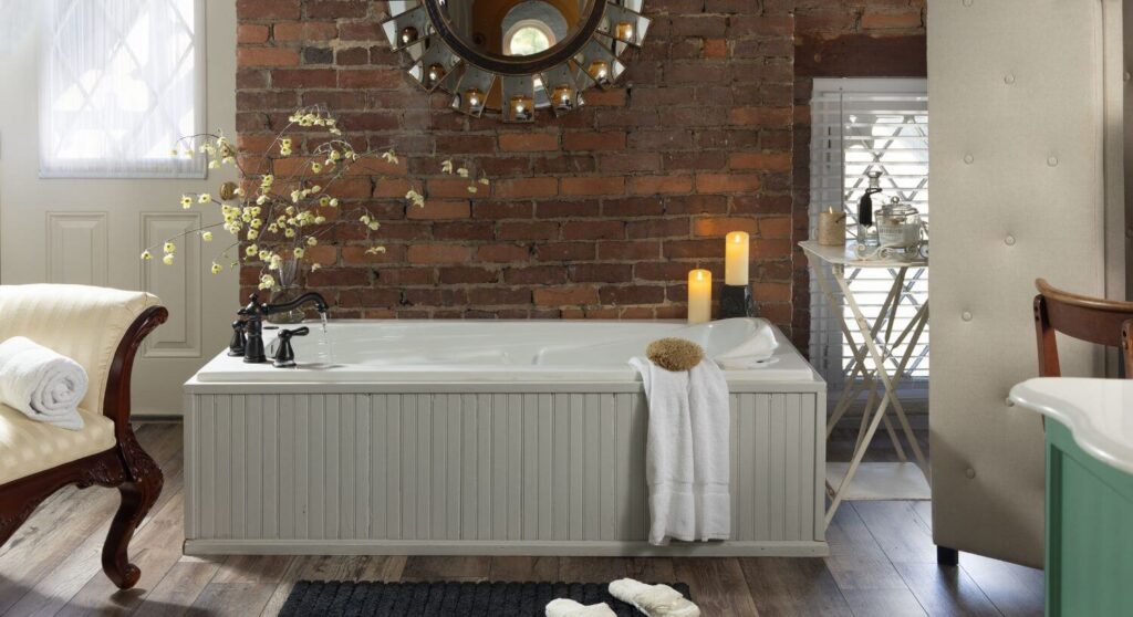 Large bathroom with tub next to red brick wall, towel hanging on edge, lit candles, wood planking flooring, slippers on rug