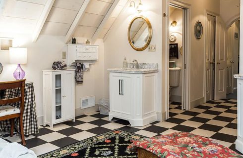 Large bathroom with white walls, black and white checked tile flooring, two white vanity cabinets with marble tops, gold-framed oval mirrors