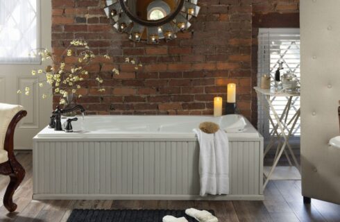 Large bathroom with tub next to red brick wall, towel hanging on edge, lit candles, wood planking flooring, slippers on rug