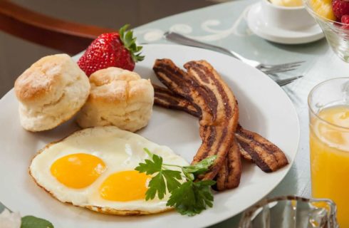 White plate with over easy eggs, slices of bacon, biscuits, large strawberry, and glass of orange juice