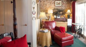 Bedroom with dark wooden four-poster canopy bed, red and white bedding, white-paneled walls, red leather chair and ottoman, and wicker side table