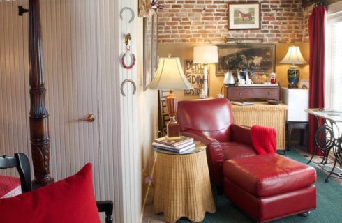 Bedroom with dark wooden four-poster canopy bed, red and white bedding, white-paneled walls, red leather chair and ottoman, and wicker side table